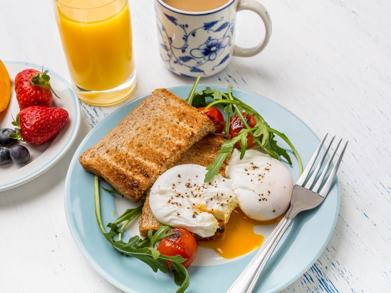 Wholegrain toast with poached eggs, tomatoes and spinach leaves on a blue plate, with orange juice, tea and berries on the side