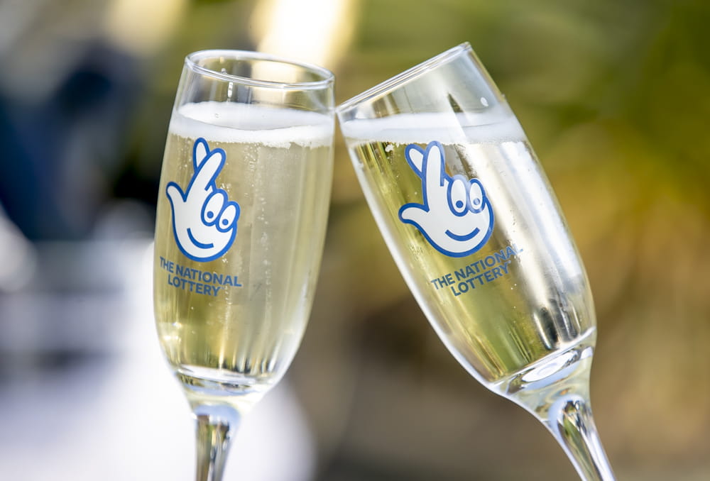 Two full champagne flutes being chinked with National Lottery logos on their sides