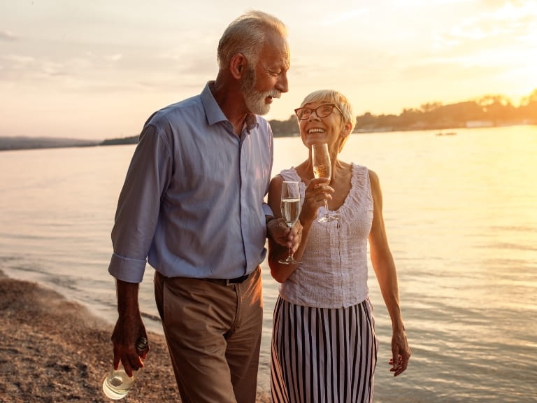 Mature couple walking along a beach drinking champagne from flutes
