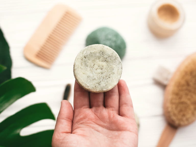 Hand holding natural solid shampoo bar on background of bamboo brush, deodorant, sponge and monstera leaves