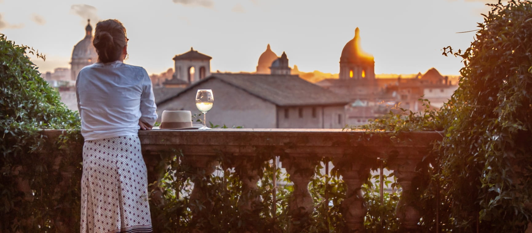A woman leaning on a wall with a glass of white wine in front of a panoramic view of the Rome cityscape