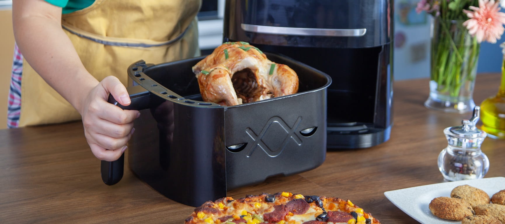 An open airfryer drawer containing a roast chicken and a pizza on the table in front of it