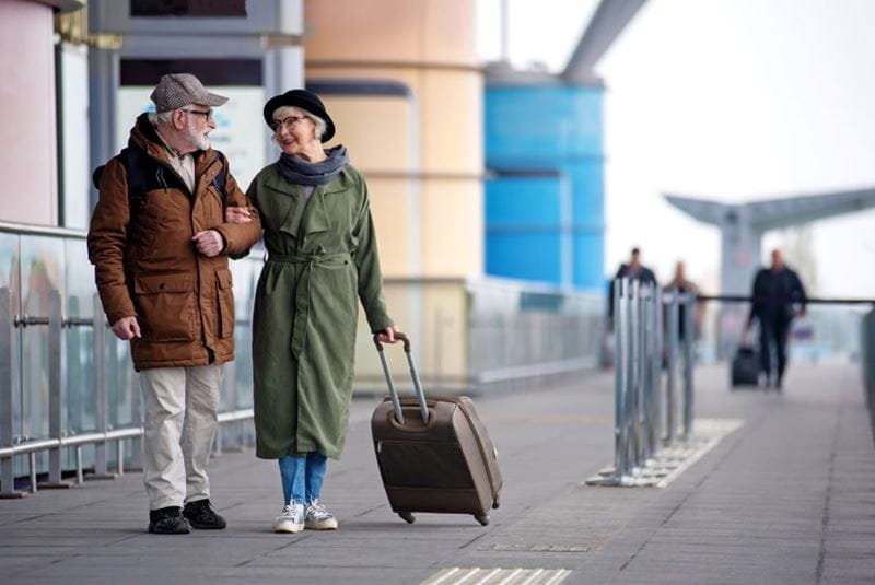 Full length of encouraged calm retired man and woman are talking to each other while standing outdoors. They are carrying suitcase while looking at each other with love