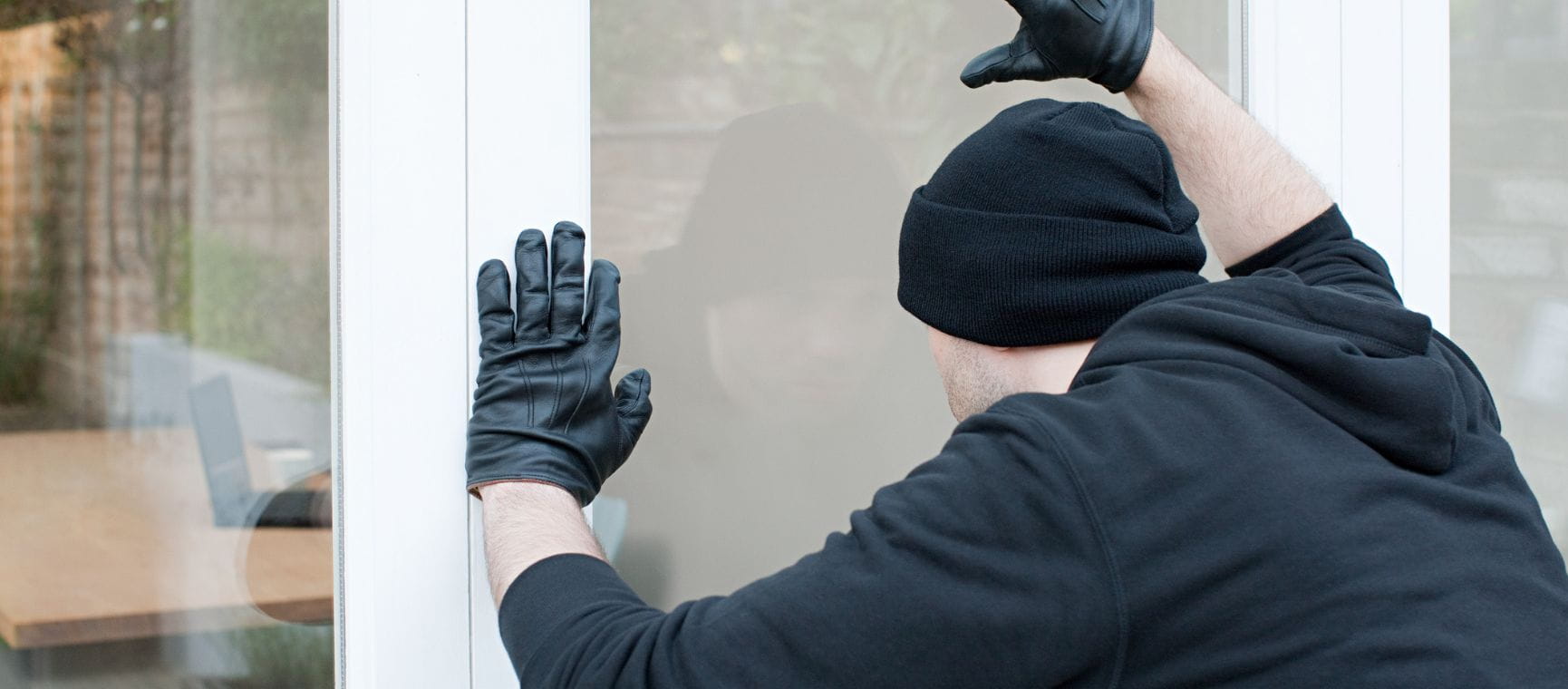 A burglar dressed in black peering through patio doors at a laptop on a table inside