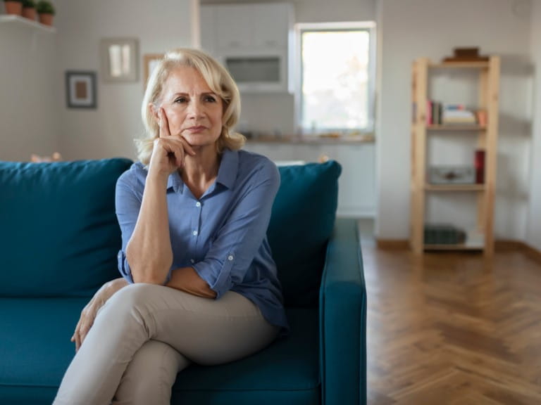 An older woman sits on a sofa looking worried