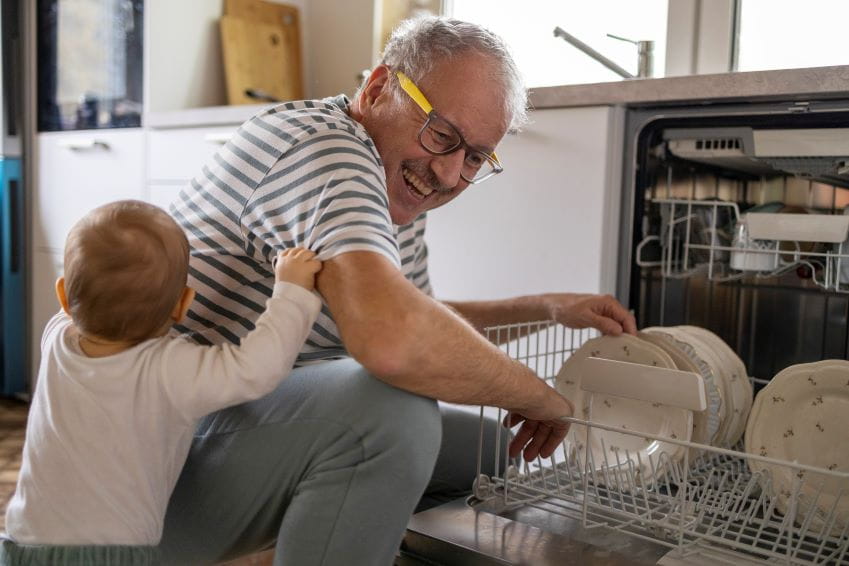 Man loading the dishwasher with a baby boy pulling at him