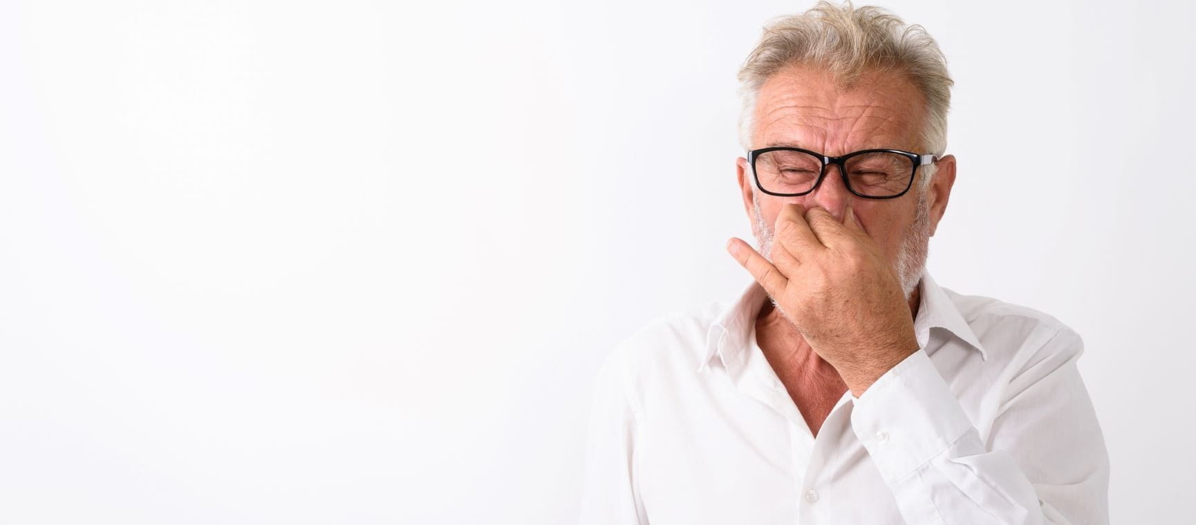 Studio shot of senior bearded man covering nose and looking disgusted while wearing eyeglasses against white background horizontal shot