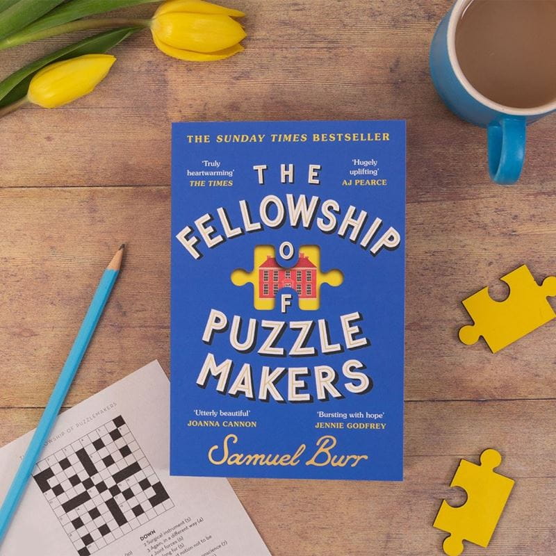 A copy of The Fellowship of Puzzle Makers on a wooden table next to puzzle pieces and a crossword