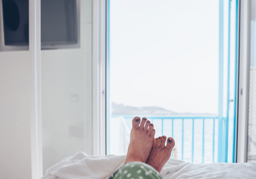 Feet poking out of bed covers in a hotel room overlooking the sea