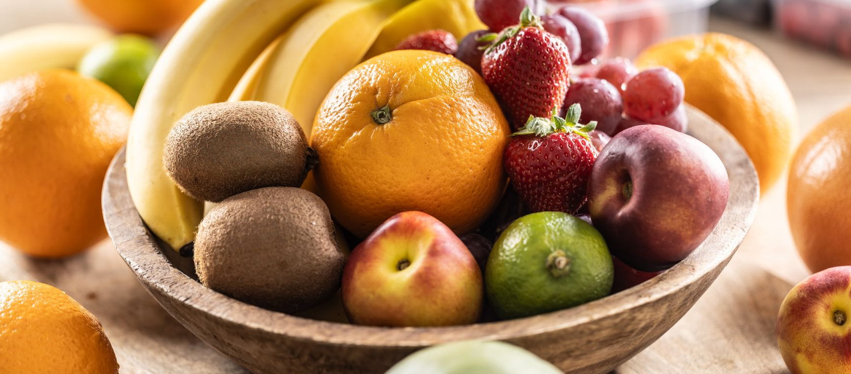 A wooden bowl full of fruit such as bananas and oranges sat on a table surrounded by more fruit
