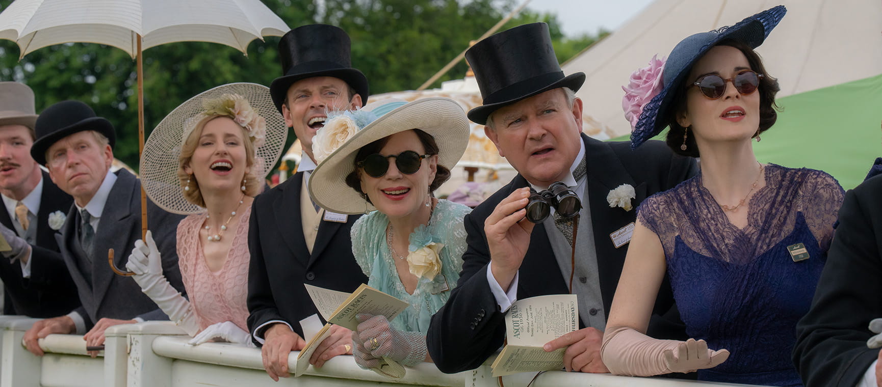 A group of people in 1930's dress including top hats and suits leaning over a barrier with looks of anticipation