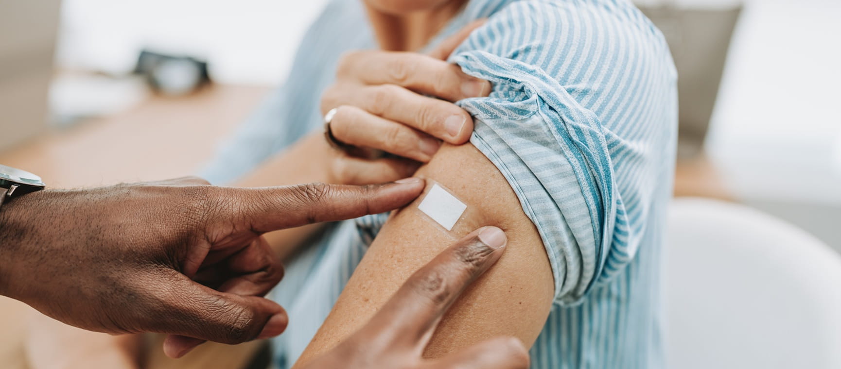 Hands placing a plaster on an upper arm with the sleeve rolled up, suggesting a vaccination