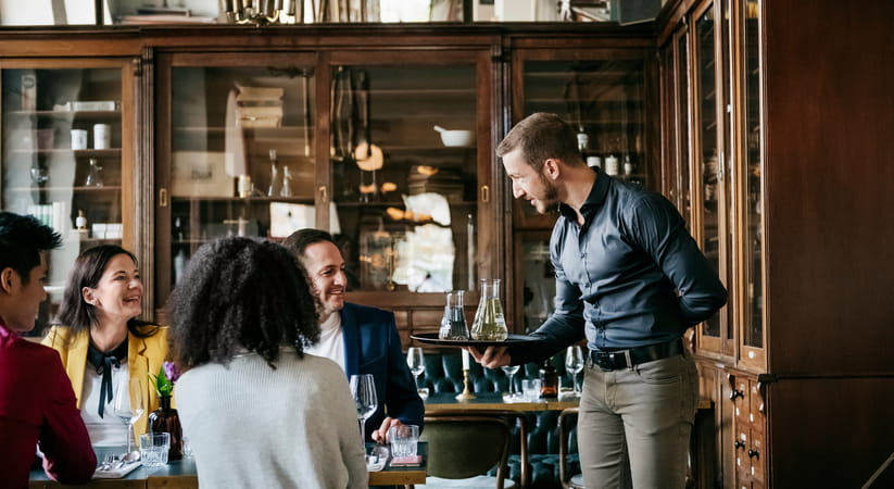 Waiter in blue shirt and grey trousers serving group of men and women in a restaurant