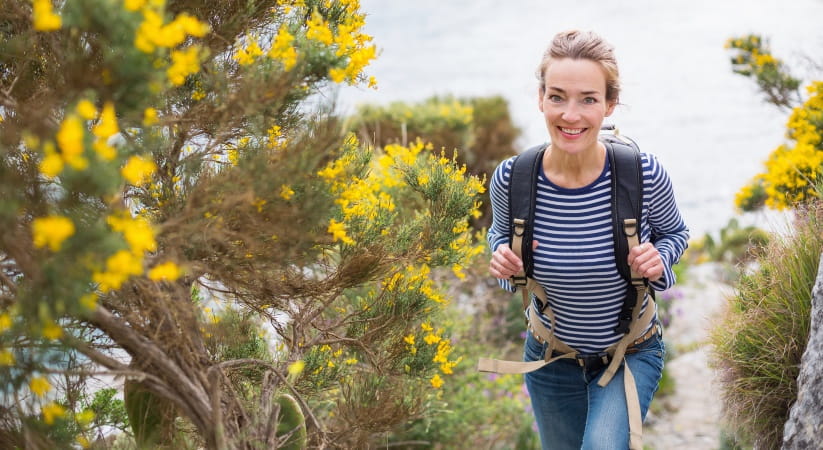 Woman in stripy top wearing rucksack walking up a coastal path and smiling