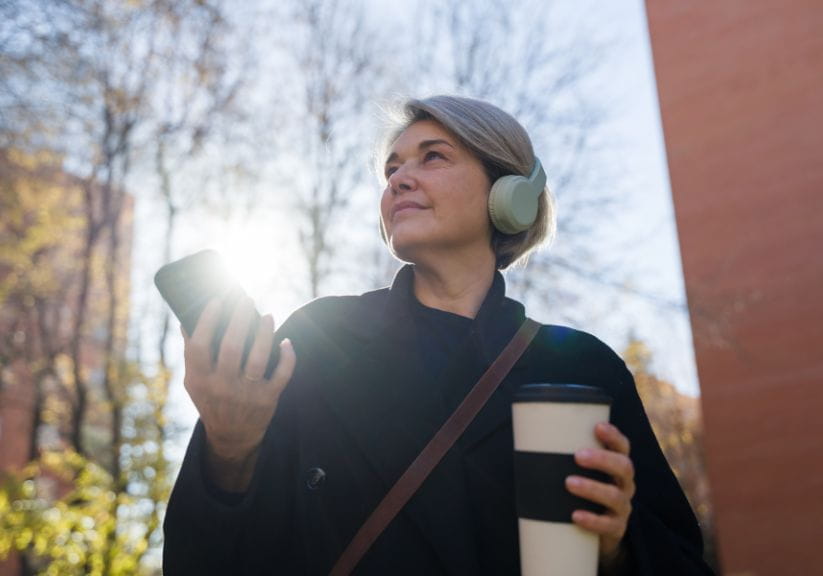 A mature woman listening to a podcast on their phone outdoors while holding a coffee
