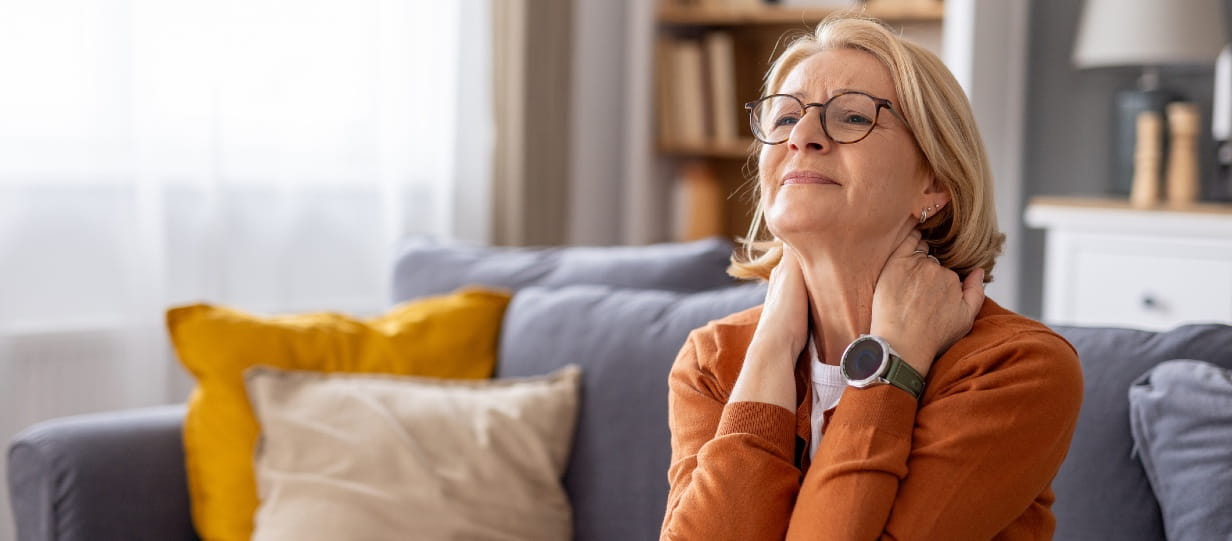 Older woman in orange top and glasses sat on sofa and holding her neck