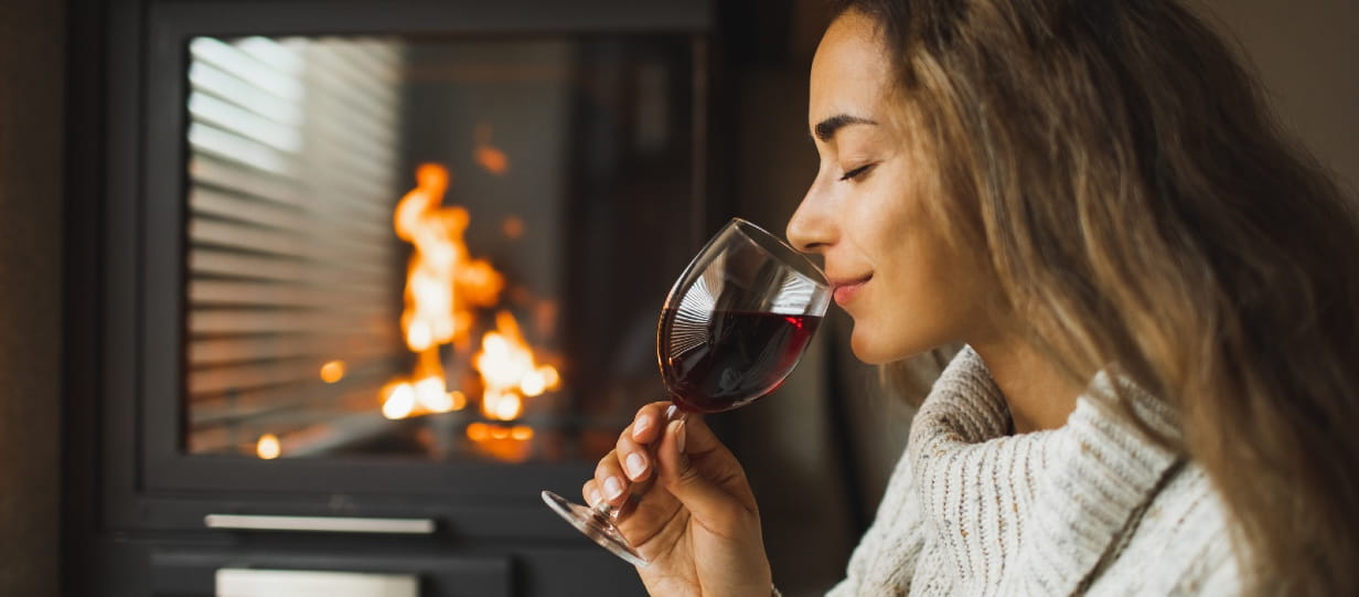 Young woman in a white jumper smelling a glass of red wine while standing next to a roaring fire
