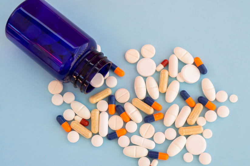A pile of pills of different sizes, forms and colours on a pastel blue background coming out of a blue jar