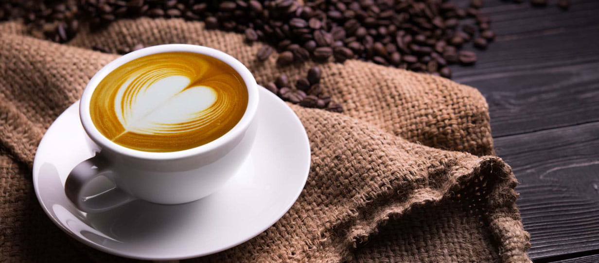 A white cup of coffee on a wooden table surrounded by coffee beans