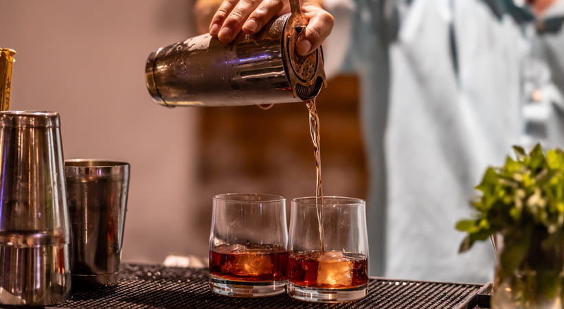Close up of a bartender in a white shirt pouring two drinks with a cocktail shaker