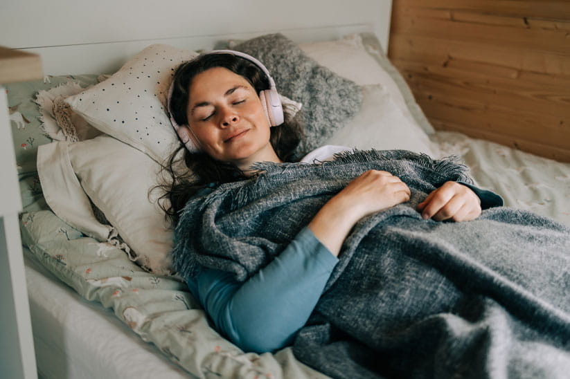 Woman relaxing in bed listening to music with headphones, covered by blanket