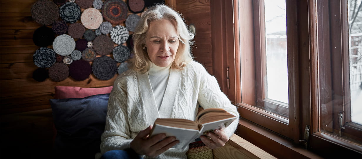 An older woman reading a book by a window showing snow outside