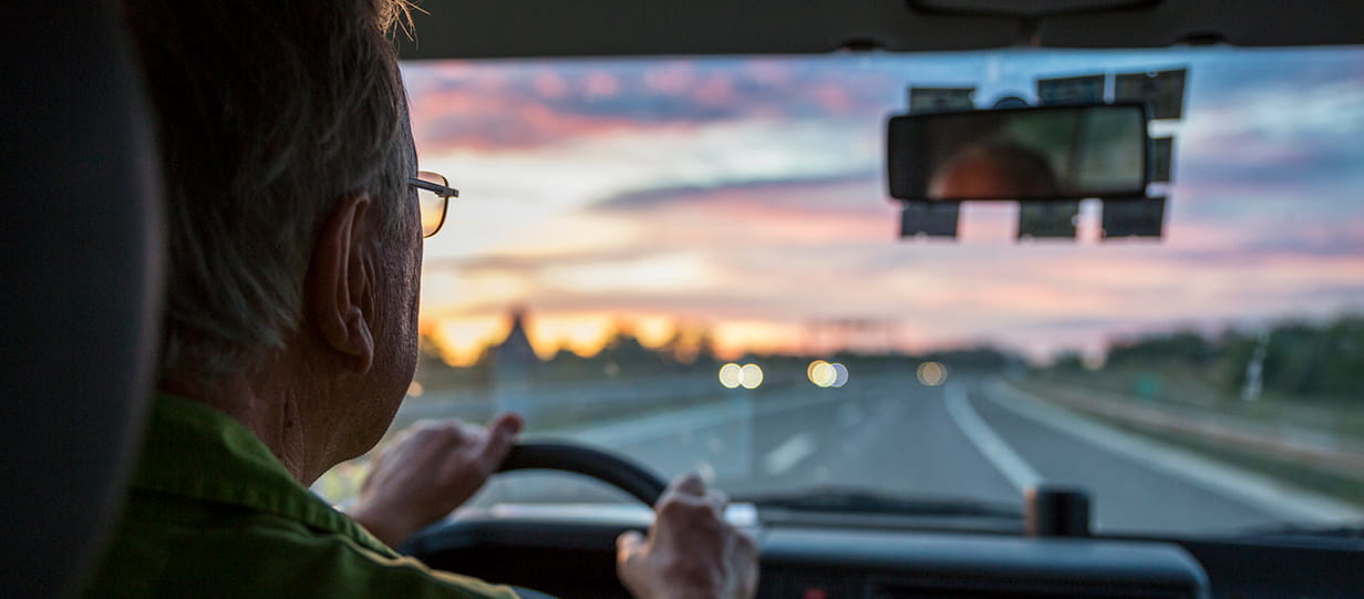 An older driver wearing glasses driving at sunset