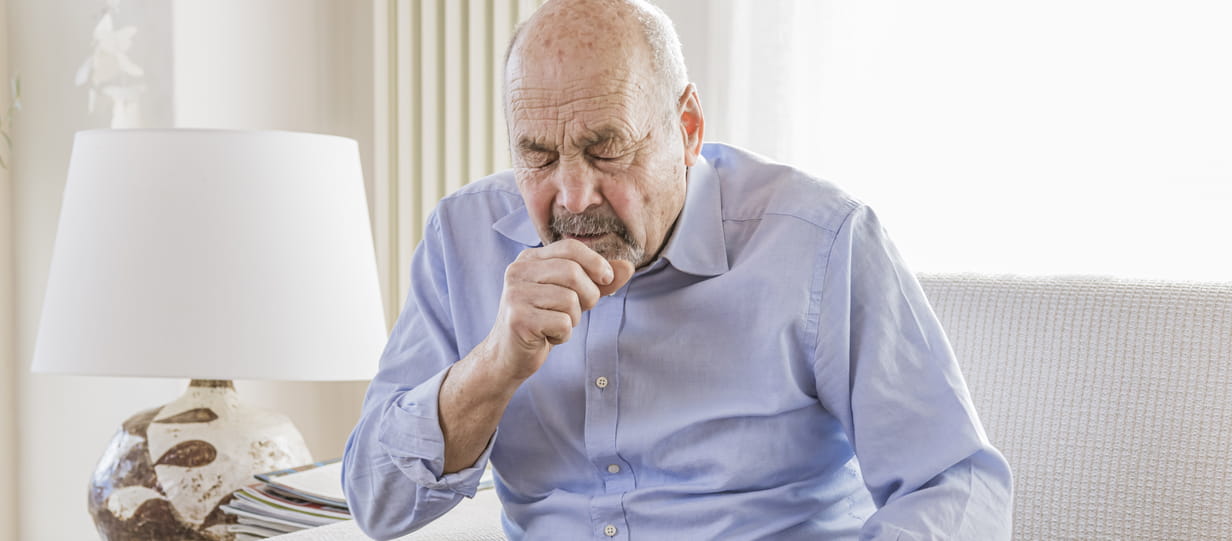 An older balding man in a blue shirt coughs while sitting on a sofa