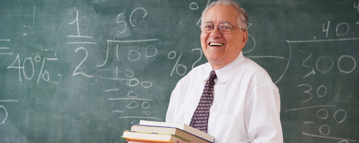 Mature Professor holding a pile of books and smiling with mathematical writing on the chalk board behind him