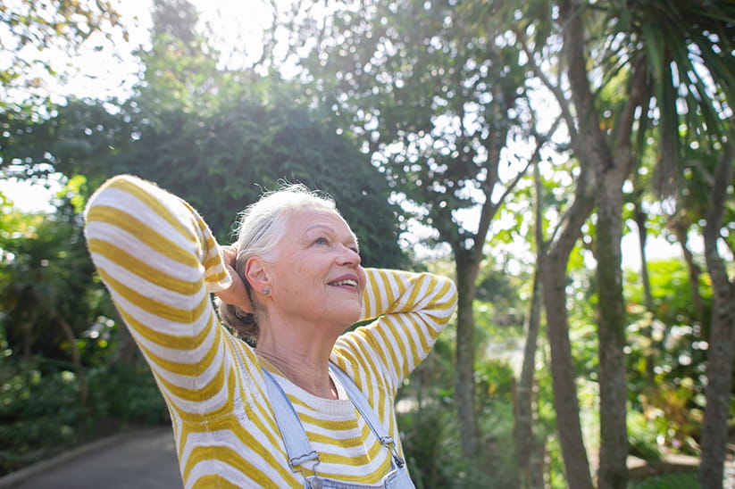 An older lady smiling in the sun in front of some trees