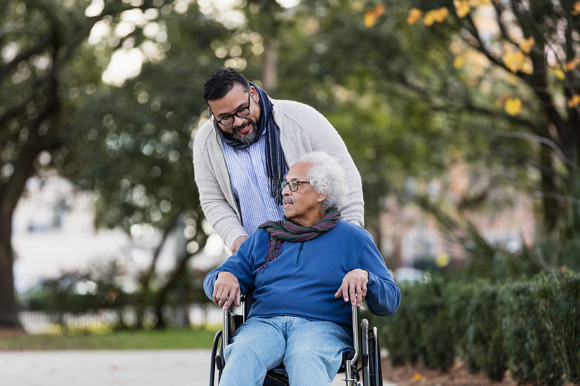 A senior Hispanic man in his 80s sitting in a wheelchair, taking a walk in the park with his adult son, an adult man in his 30s.