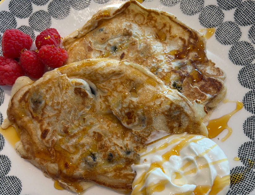 Two golden pancakes made with blueberries using Jamie Oliver's recipe on a plate with raspberries and cream garnish