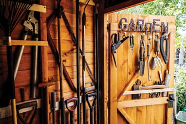 Inside view with open door of organised shed filled with rake, shovels, trimmer, saw, and other hand tools required for maintenance of well tended garden.