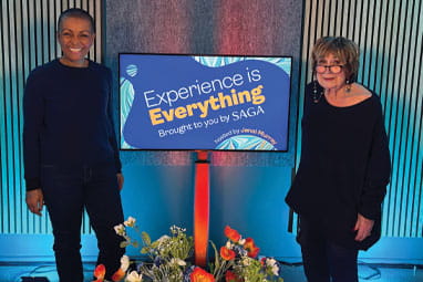 Adjoa Andoh (left) and and Jenni Murray (right) standing on either side of a screen displaying the Experience is Everything podcast logo