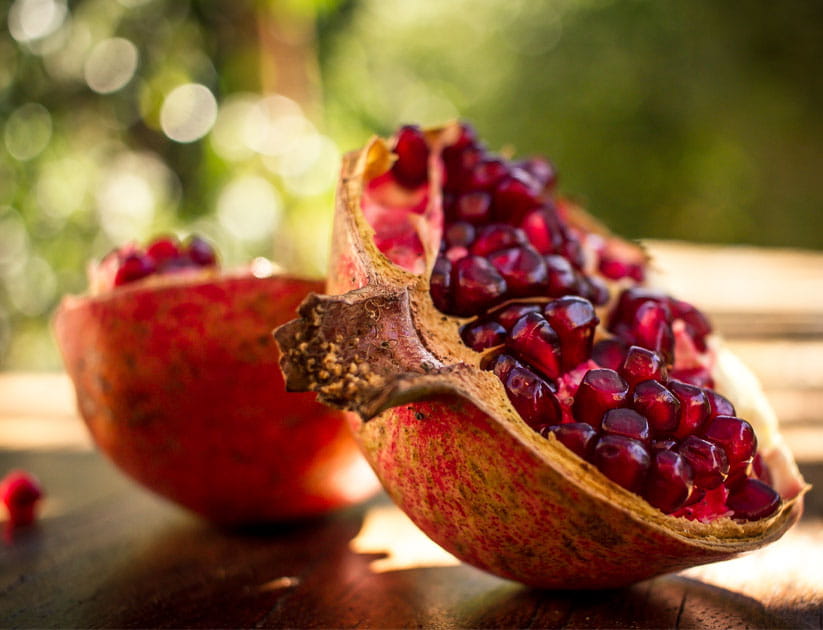 Ruby Red Pomegranate split open on a wooden table with green foliage as a blurred background.