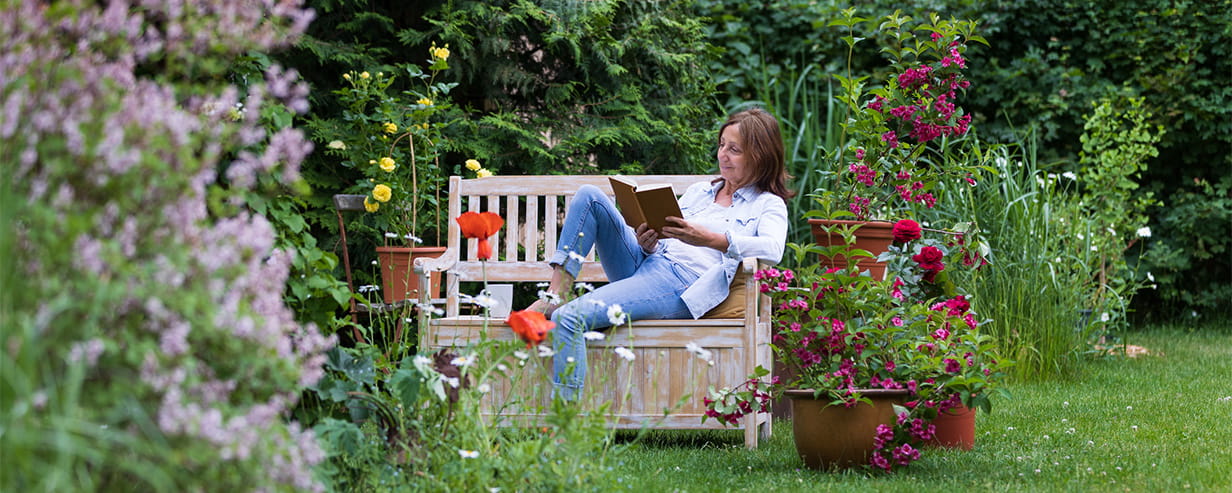 An older woman reading her book whilst sat on a bench in a garden, surrounded by flowers