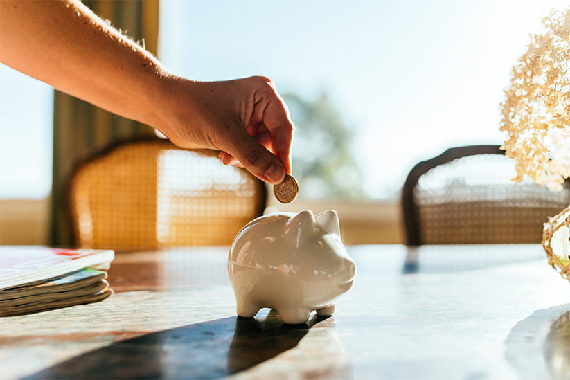 A woman placing a coin in a white piggybank that's sitting on a wooden table in the sunshine