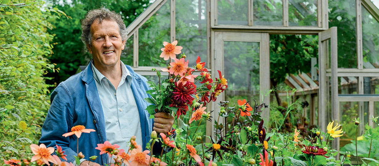 Monty Don smiles as he stands in front of a greenhouse in a flower garden