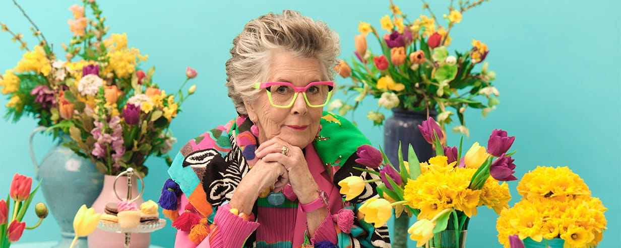 Prue Leith sitting surrounded by colourful flowers, in front of a light blue wall