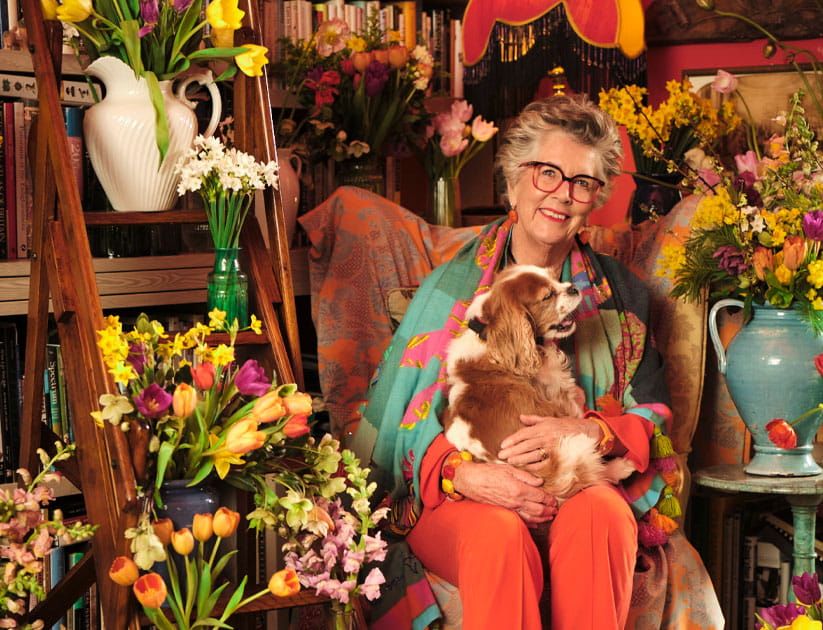 Prue Leith with one of her spaniels on her lap surrounded by British spring flowers and books