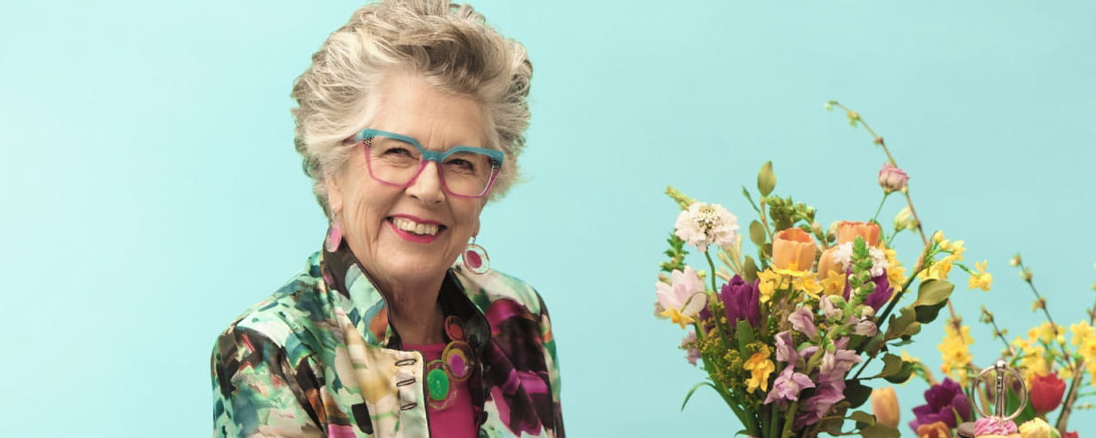 Head and shoulders photo of Prue Leith against a light blue background with spring flowers next to her