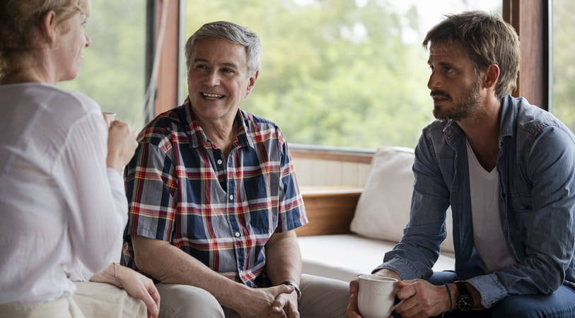 An adult man in a blue shirt and white T-shirt sits and talks with his mother and father