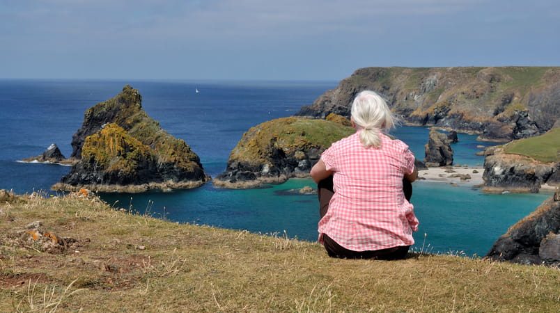 Rear view of an older woman in a pink shirt sitting on a clifftop in Cornwall and looking out to sea