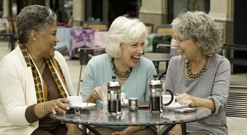 Three older women sit around a table at an outdoor cafe laughing