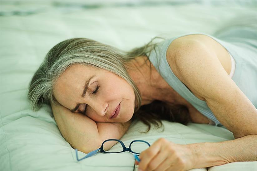 Mature woman with long grey hair and blue glasses taking an afternoon nap on a green bed.