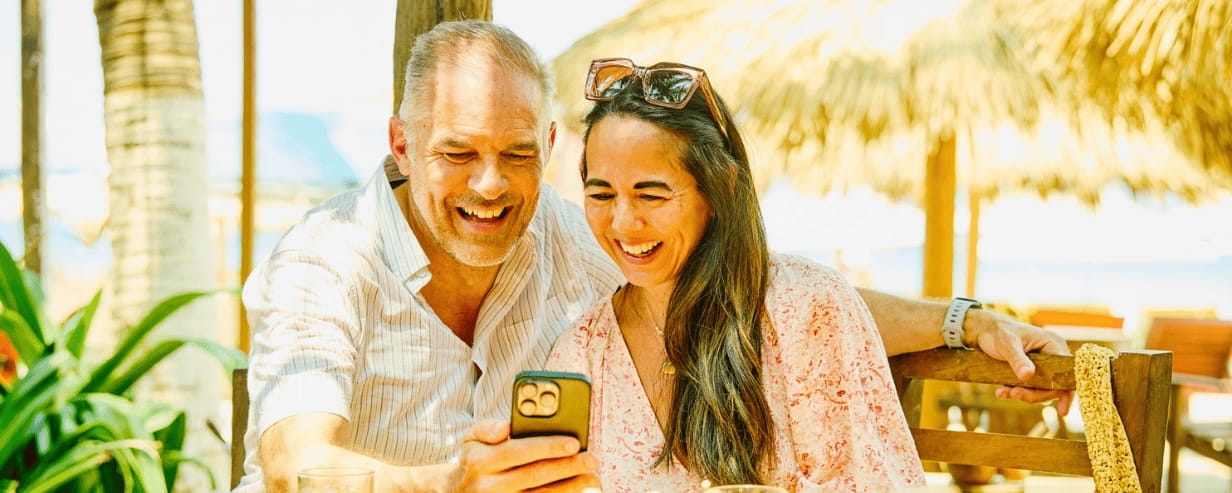 A couple smiling into a mobile phone at a beach cafe