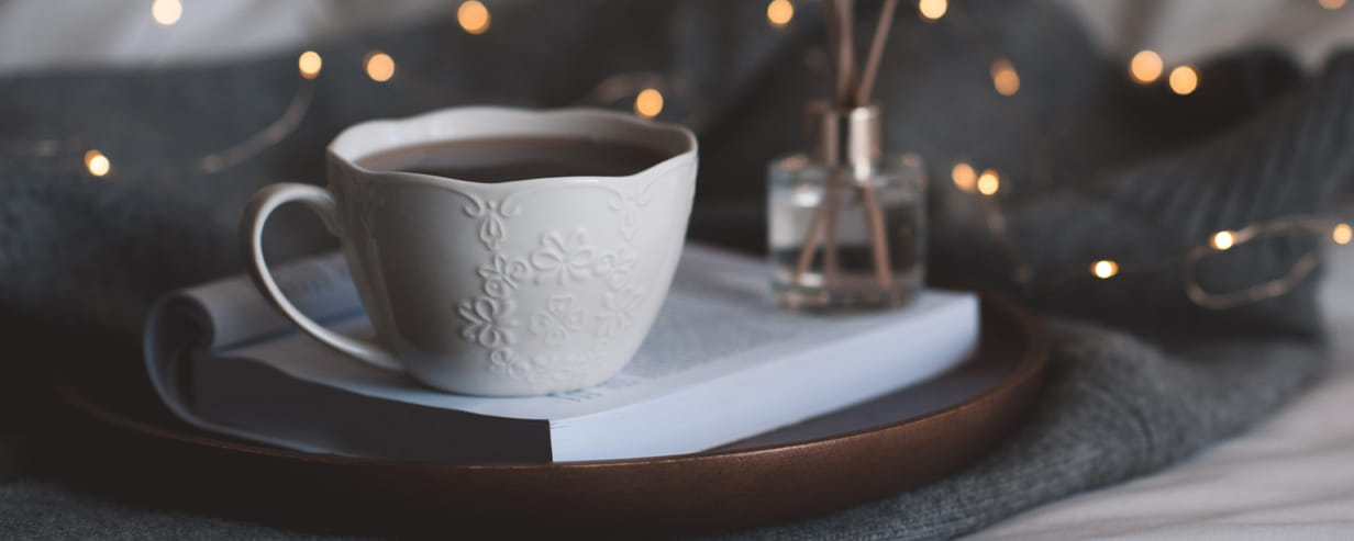 Cup of coffee on open paper book with a reed diffuser on wooden tray with led sting lights in bed over knit cloth closeup