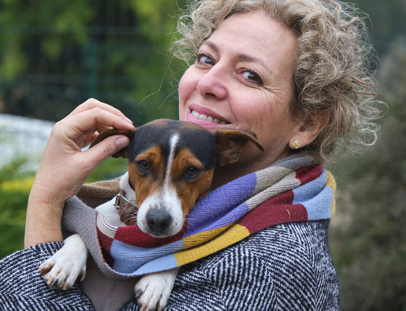 Mature woman smiles while holding her small dog, wrapped in a colorful scarf, in a garden setting
