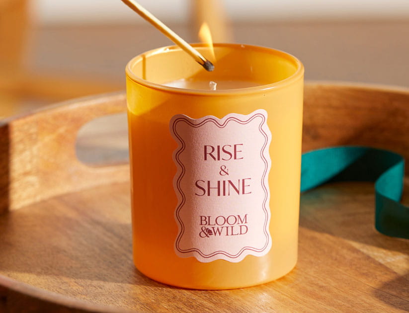 A scented candle in a light orange glass with a lit match about to light the wick. text reads: Rise & Shine Bloom & Wild