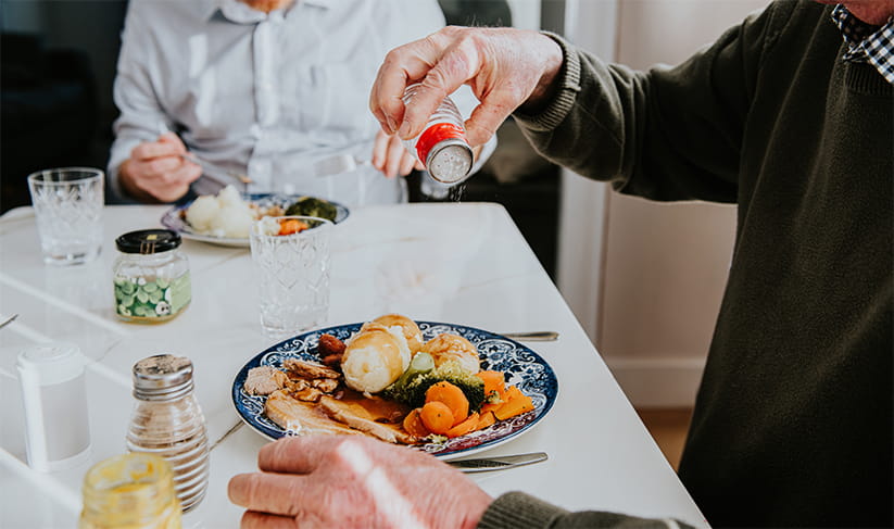 A man pours salt onto his plate filled with vegetables and meat while dining with another individual in a warm, inviting room, creating a comforting atmosphere